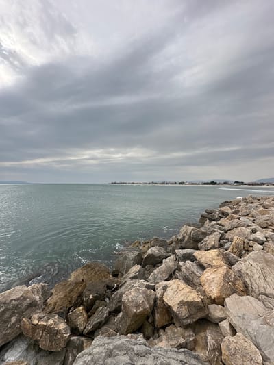 This image depicts a coastal scene in Limenas Avdiron, Greece, under an overcast sky. The foreground and right side are dominated by a rocky breakwater or natural shoreline composed of large, irregularly shaped boulders in varying shades of grey, brown, and tan. The rocks meet the calm, greenish-blue sea, which occupies the midground and left portion of the image. Small whitecaps are visible where the water meets the rocks. In the distant background, along the horizon, a low-lying coastline features what appears to be a small coastal settlement or town, marked by visible buildings and structures. Further inland, faint outlines of hills or mountains are discernible. The sky is entirely covered with thick, grey clouds, indicating overcast weather conditions. No individuals, animals, or specific objects indicating human activity (beyond the distant settlement) are present in the immediate scene.