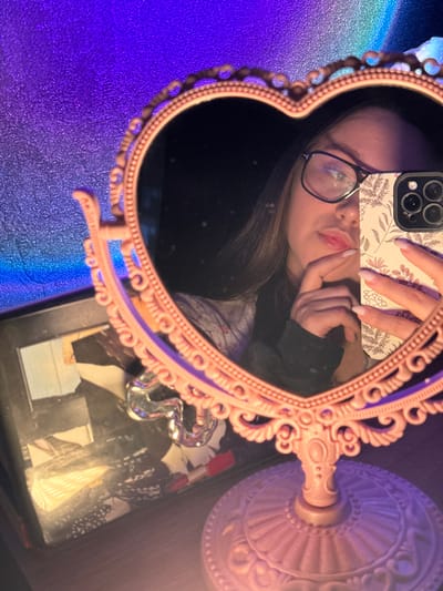 The image depicts a mirror selfie taken by a female in what appears to be a dimly lit indoor setting, attributed to the city of Prikubanskii, Russia. The central object is an ornate, pink, heart-shaped vanity mirror with a decorative pedestal stand, positioned on a flat surface. The reflection in the mirror shows a female, visible from the upper chest, with long brown hair, wearing black-framed eyeglasses. She is holding a light-colored smartphone with a floral-patterned case and a visible triple camera array in her left hand, which partially obstructs her face. Her right hand is posed near her chin or lower lip, with light-colored fingernail polish visible. She has slightly pursed lips and is wearing a light-colored top beneath a dark long-sleeved garment. The background outside the mirror's frame on the left features a textured wall illuminated by a gradient of vibrant purple to blue light. To the lower left, a dark rectangular object, possibly a book or box, with reflective details, is visible next to a clear, abstract decorative item.