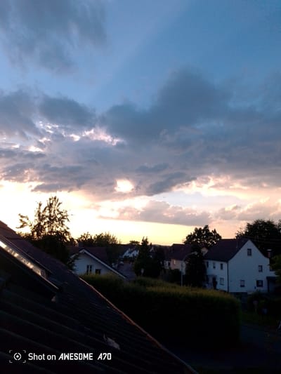 A sunset is captured over a residential area in Lohmar, Germany. The sky is filled with dramatic, multicolored clouds, with streaks of yellow and pink light breaking through the darker gray masses. In the foreground, the dark, angled roof of a building with a skylight dominates the lower left of the frame. Beyond the roofline, silhouetted trees and the rooftops of houses are visible, illuminated by the fading sunlight. The houses are of typical European design, with tiled roofs and white or light-colored facades. The overall impression is of a tranquil evening scene. The image also contains a watermark in the lower left corner that reads "Shot on AWESOME A70".