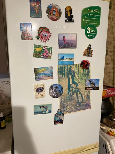 The image shows a refrigerator adorned with numerous magnets and stickers. The refrigerator is white and is located in a kitchen environment. The magnets and stickers feature various designs, including cartoon characters, landmarks, and scenic views. There are also some text-based stickers, some of which are in Russian and appear to be advertisements or labels. One prominent sticker is a green oval shape with text and a logo that seems to indicate a product guarantee. The overall impression is of a decorated and personalized refrigerator, typical of a home setting. The specific location in the city of Маринопіль, Ukraine, is not discernible from the image.