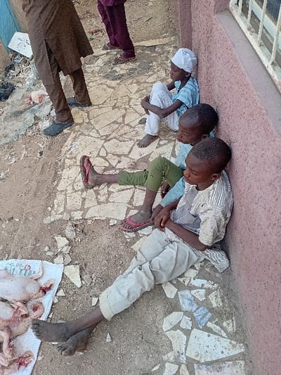 An outdoor scene in Sharada, Nigeria, features three young males seated on a dirt and broken tile surface. The boy in the foreground wears a striped long-sleeved shirt and light beige trousers, his bare feet extended, leaning against a reddish-brown stucco wall. To his left, a second boy wears a light blue top, green trousers, and pink flip-flops. A third boy, further back, wears a light blue top, white trousers, and a white cap, sitting with his knees drawn up. All three are positioned in various states of repose. To the immediate left of the seated individuals, several pieces of raw, plucked poultry, displaying visible blood, are arranged on white material directly on the ground. In the background, the lower legs of two adults are partially visible; one in brown trousers and dark shoes, and another in purple trousers and sandals. The reddish-brown wall behind the boys includes a partial view of a white-barred window in the upper right. The ground throughout the scene is uneven and contains general debris, including dark plastic bags.