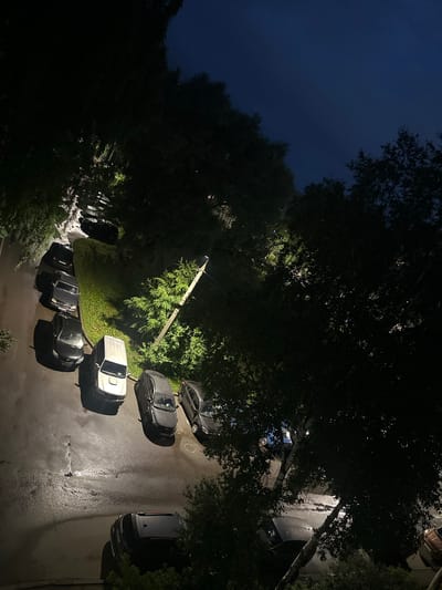 The image depicts a nighttime scene of a parking lot with several cars parked on wet asphalt. The lighting suggests a streetlamp or multiple light sources illuminating the area. The cars appear to be parked in a residential area, with trees and foliage visible on the sides and overhanging the parking lot. One car is white, while others are dark colors like black and gray. The sky is dark blue, indicating it is nighttime. The overall impression is of a quiet, somewhat damp evening in an urban or suburban setting. The specific location in the city of Rybinsk, Russia, cannot be determined solely from the visual information provided in the image.