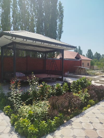 The image depicts an outdoor scene in a residential area, possibly a backyard, in the village of Жаңы-Айыл, Kyrgyzstan. In the foreground, a paved walkway with a geometric pattern leads to a lush garden filled with various plants and flowers, including roses. Behind the garden, a covered patio or gazebo stands, featuring a white corrugated roof supported by dark metal posts. The gazebo encloses a raised wooden platform, with a patterned rug and a pillow visible. A ladder leans against the brick wall behind the gazebo. Beyond the gazebo, a red brick wall encloses the property, and further back, a two-story house with a reddish-brown tiled roof is visible. Tall trees line the perimeter of the property, and the sky is a clear, bright blue. The overall impression is one of a well-maintained outdoor living space in a rural or suburban setting.