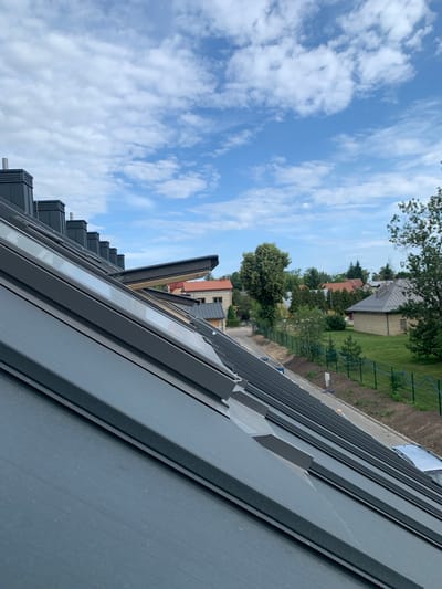 The image shows a close-up view of a dark gray metal roof with multiple skylights, some of which are open. In the background, several houses with red and gray roofs are visible, surrounded by green trees and lawns. A dark green fence runs along the property line. The sky is blue with scattered white clouds. The scene appears to be a residential area. The location is Białystok, Poland.