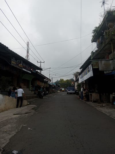 The image presents a street scene in the city of Houaythat, Laos, viewed under an overcast, light-grey sky. The perspective looks down a relatively narrow, asphalt-paved street flanked by commercial and residential structures.
On the left side of the street, a series of single-story, open-front shops with dark, corrugated metal roofs extends into the mid-ground. Merchandise, including packaged snacks and various hanging items, is displayed at the storefronts. In the immediate foreground, a concrete sidewalk or raised ledge serves as a seating area for three individuals. One male, wearing a white short-sleeved shirt and dark trousers, stands with his back partially to the viewer, descending a step from the ledge. Two motorcycles are parked curb-side further down the street on this side. Above the rooftops in the mid-distance, a tall, red and white lattice-style cellular or radio tower is prominently visible. Dense green foliage and hills rise in the far background.
On the right side of the street, the structures are more varied, including a multi-story building with weathered concrete facades and green vegetation growing on ledges. An antenna array is mounted on the roof of this building, and clothing is observed hanging from a balcony. Adjacent to it, an open-front shop is visible, where two females are positioned; one is seated while the other stands, both appearing to interact with goods inside. A large white banner with text is affixed to the shop's exterior, alongside smaller signs. Further down the street, a dark blue SUV is parked facing away from the viewer.
Numerous utility poles and a dense network of overhead power and communication lines crisscross above the street and buildings. The asphalt road surface exhibits minor cracks, and scattered debris is present on both the road and the uneven concrete sidewalks. The overall environment suggests a bustling commercial thoroughfare.