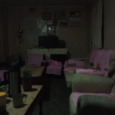 A dimly lit room holds a mix of furniture. A table in the foreground displays a thermos, a bottle with dark liquid, a mug, and a jar. The background reveals a television set atop a white-covered table with various decorative items above. Seating, including sofas and chairs, are draped with pink cloths. The room has a lived-in, cluttered appearance, adding to the sense of a cozy, inhabited space.