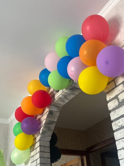 A garland of multicolored balloons is attached to a white ceiling and a brick archway in an interior space. The balloons are arranged in a curve, with colors including red, orange, yellow, green, blue, pink, and purple. Some balloons are partially obscured by others. The brick archway has a light grey tone and a rough texture. Below the archway, a dark, patterned wallpaper is visible on the wall, and wooden door frames are present. The lighting suggests an indoor setting, likely a home or event space.