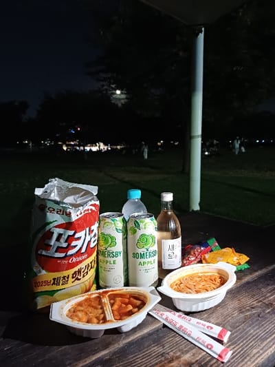 The image depicts a nighttime scene in a public park within 영등포구, South Korea. A collection of food and beverages is arranged on a dark wooden surface, likely a picnic table, in the foreground.
**Objects:**
* **Food:** One partially opened bag of "Orion Pocachip Onion" potato chips, a white disposable container divided into two sections, holding a reddish-orange stew (likely tteokbokki) and a lighter-colored, similar dish. Another white oval disposable container holds red-orange noodles. Several small, colorful, partially obscured snack pouches are positioned behind the noodle dish.
* **Beverages:** Two green cans of "Somersby Apple" cider, one clear plastic bottle of water with a light blue cap, and one clear glass bottle with a white label reading "새로" (Saero), containing a light-colored liquid (likely soju).
* **Utensils:** Two sets of individually wrapped disposable chopsticks, featuring red and white packaging with Korean text, are placed beside the noodle container.
* **Environment:** The background is dark, indicating night. A large, grassy park area is visible, with dark silhouettes of trees against the sky. A bright, nearly full moon is discernible through the trees. Scattered, blurred figures of people are present in the distant park, suggesting other individuals are occupying the space. A prominent, light-colored vertical pole or pillar stands on the right side of the frame, extending from the bottom edge upwards.
**Actions/Interactions:**
The arrangement of food and drink implies preparation for or ongoing consumption by one or more individuals, although no people are explicitly interacting with the items in the foreground. The distant figures suggest general recreational activity in the park.
**Location:**
The scene is set in a public park in 영등포구, South Korea.