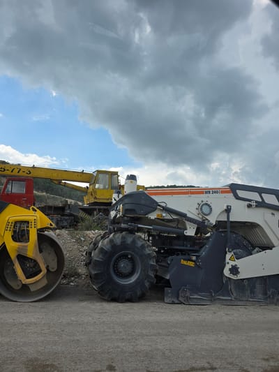 The image captures a scene at the Gara Oreshets, Bulgaria, featuring heavy construction machinery. In the foreground, a yellow road roller is parked on a gravel surface. To its right, a large white and gray asphalt milling machine is visible, with its heavy-duty wheels and cutting drum prominent. Behind these machines, a yellow crane with the partial number "5-17-3" on its boom is positioned. A red truck cab is also present to the left of the crane. The background consists of a hillside covered with vegetation, under a cloudy sky.