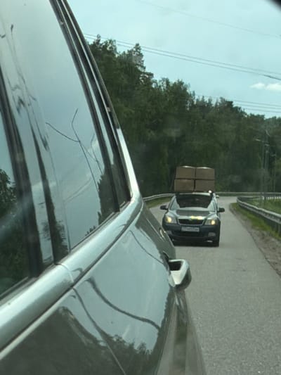 The image shows a car driving on a paved road. Another car is visible ahead, carrying several cardboard boxes on its roof. The background features a forest area and some power lines. The photo was taken from inside a car. There is no evidence to suggest the location is Plastik, Russia.