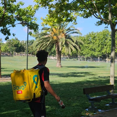 The image shows a person from behind, wearing a yellow delivery backpack with the "Glovo" logo and "FLiiTS" text. The person is walking through a park with green grass, trees, and sprinklers in operation, suggesting it's daytime. In the foreground, a wooden bench is visible, wet with water, and to the right, a concrete pole stands. The person is holding a partially consumed bottle in their right hand. The scene is set in the Parque del Agua Luis Buñuel in Zaragoza, Spain, as indicated by the specific design of the yellow delivery bag, which is characteristic of Glovo deliveries in that region. The backdrop features more trees and some buildings in the distance, under a clear blue sky.
