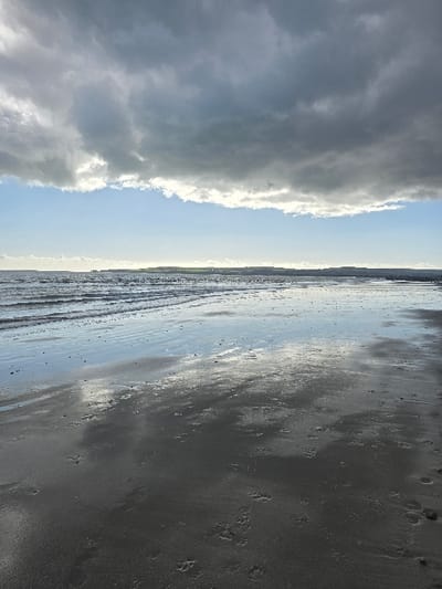 This image depicts a coastal scene on a beach in Poole, United Kingdom, under an overcast sky. The upper two-thirds of the frame are dominated by a dense formation of dark grey stratus clouds. A horizontal band of clear, light blue sky is visible below the main cloud mass, illuminated by what appears to be concealed sunlight, creating a bright glow along the cloud's lower edge.
The horizon line, positioned approximately one-third up from the bottom of the image, features a distant landmass identified as a coastline. This coastline appears to include elevated terrain, possibly cliffs or hills, with visible patches of green vegetation and some indistinct structures. Faint, dark specks are discernible on the water surface in the mid-distance, likely distant objects or craft.
The sea occupies the middle ground, exhibiting gentle wave action near the shoreline. The water's surface reflects the sky, particularly the brighter blue and white sections.
The foreground consists of a wide expanse of wet, dark sand. The moisture in the sand causes it to be highly reflective, mirroring the cloud formations and the bright strip of sky above. Numerous imprints are visible in the wet sand: a concentration of human footprints, varying in size, and multiple distinct paw prints consistent with a canine. No individuals or animals are present in the immediate foreground or mid-ground. The overall lighting suggests an overcast daytime environment, with indirect sunlight providing illumination and creating reflections.