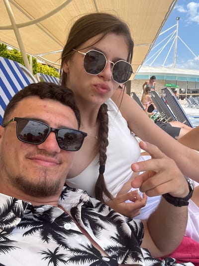 This close-up, rotated selfie features two individuals, a man and a woman, relaxing outdoors in what appears to be a resort or beach setting in Selçuk, Turkey. The image is oriented with the horizon running vertically.
In the foreground, the woman is positioned above the man, resting her arm on him. She has long brown hair, styled in a braid tied with a black band, and wears gold-rimmed hexagonal sunglasses. She is dressed in a white ribbed tank top and has a delicate necklace around her neck. Her lips are slightly pursed, giving her a playful expression, and her right hand is visible in a gesture with fingers extended.
The man is situated below the woman, wearing black rectangular sunglasses. He has short brown hair and a beard and is dressed in a white shirt with a black palm tree pattern. A black watch is visible on his left wrist. His facial expression is largely obscured by his sunglasses.
The background shows a bright, sunny daytime scene with a clear blue sky. A light brown or tan canopy structure provides shade. Numerous lounge chairs are visible, some with blue and white stripes, suggesting a resort or beach area. Other people are seen relaxing on these chairs in the distance, including one person in a blue swimsuit. A tall white pole-like structure with supporting wires is silhouetted against the sky. The overall atmosphere conveys a relaxed holiday or vacation mood under bright daylight. There is no visible text in the image.