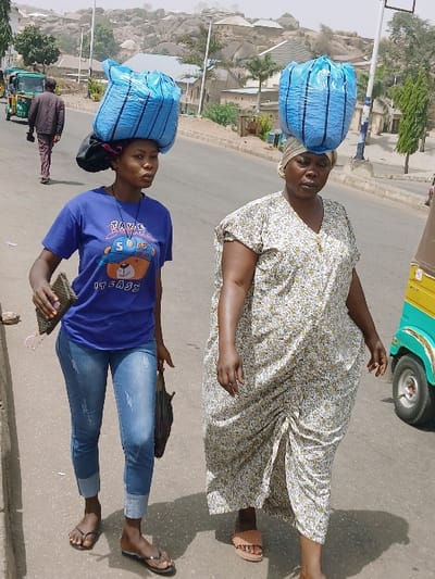 The image depicts an outdoor street scene in Kadoma, Zimbabwe, under bright daylight. Two women are prominently featured walking right-to-left along a paved road's edge. The woman on the left, wearing a blue t-shirt with a graphic, blue jeans, and black flip-flops, carries a small dark handbag and a patterned wallet; a large, light blue bundle is balanced on her head. The woman on the right, dressed in a long, patterned cream and green dress with a light headwrap and tan sandals, also balances a similar large, light blue bundle on her head. Both women possess dark skin tones. In the background, a man in a dark shirt and maroon trousers walks away from the camera on the road, past a green and yellow three-wheeled vehicle (auto-rickshaw/tuk-tuk). The background features rocky hills, scattered residential buildings, and sparse vegetation, indicating a semi-urban or rural-urban fringe environment. Another section of a yellow and green three-wheeled vehicle is partially visible on the far right. The ground is asphalt, with a lighter-colored sidewalk or shoulder to the left.