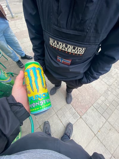 An overhead shot captures a casual outdoor scene in Vetrino, Bulgaria, with at least three individuals standing on a paved surface. The ground is a patchwork of light grey and reddish square tiles. In the foreground, a hand holds a vibrant yellow Monster Energy drink can, emblazoned with "MONSTER PUNCH + ENERGY" and "RIO PUNCH" in bold green and blue lettering.
To the left, a person is seen from the chest down, wearing a dark blue jacket with a prominent "NAPAPIJRI graphic" logo and a Norwegian flag patch on the back. They are dressed in black pants and dark grey or black sneakers. Further to the bottom left, another person wears light blue jeans and black sneakers. On the right side of the frame, the legs and feet of a third individual are visible, clad in dark pants and dark grey sneakers with distinct striped patterns.
A green metal railing is partially visible at the bottom of the frame, with what appears to be another green drink can resting on it, beneath the Monster can. The lighting suggests it is daytime, possibly overcast, as there are no harsh shadows. The overall impression is a casual gathering of friends.