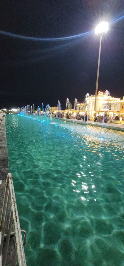 This image depicts an outdoor scene at night, likely in the city of Satkak, Uzbekistan, featuring a long, narrow canal or waterway lined with fountains. The water is illuminated with blue and white lights, creating a vibrant and dynamic effect. Along the far side of the waterway, a building with ornate architecture and illuminated windows is visible, suggesting it might be a commercial establishment or a public building. A bright floodlight on a tall pole is positioned on the right side of the frame, casting light over the scene and illuminating the night sky. Several people are gathered along the edge of the waterway, some appearing to be seated and others standing, suggesting a public gathering or recreational area. The overall atmosphere is one of nighttime entertainment and public space utilization.