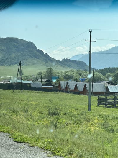 The image depicts a rural scene in Tuekta, Russia. A grassy field with scattered yellow wildflowers dominates the foreground. In the middle ground, a small village is visible, characterized by wooden houses with gabled roofs, some of which are painted red, and others with gray metal roofs. A wooden fence, constructed from rough-hewn logs, stands in the right portion of the frame. Several utility poles with electrical wires stretch across the scene. In the background, lush green trees form a dense line, and beyond them, rolling, grass-covered hills rise towards a hazy, light blue sky. The overall impression is of a quiet, pastoral landscape.