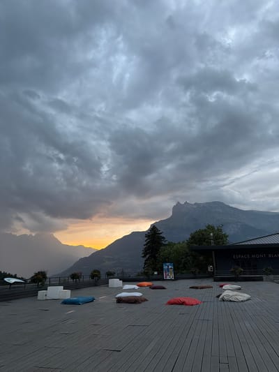 The image depicts a spacious wooden deck overlooking a mountainous landscape under a dramatic, cloudy sky. The setting sun casts a warm, orange glow on the horizon, silhouetting distant mountain peaks and creating a moody atmosphere. The deck is furnished with several large, colorful floor cushions and some modern, white seating blocks, suggesting an outdoor seating or relaxation area. A building with the text "ESPACE MONT BLANC" is visible on the right side of the frame, identifying the location as the Mont Blanc Space in Saint-Gervais-les-Bains, France. The surrounding environment includes lush green trees and the imposing grandeur of the Alps. There are no people visible in the frame.