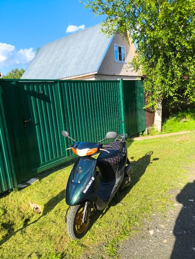 An outdoor, eye-level, medium shot captures a dark green Suzuki scooter parked on a grassy area next to a tall, dark green corrugated metal fence. The fence has a single metal handle and the number "107" is visible on it. Behind the fence, a light-colored, two-story house with a silver metal roof and a single window is visible. A large tree with green leaves is situated to the right of the house, partially obscuring it and casting shadows on the ground. The sky is blue with scattered white clouds, and the sun is shining. The ground is covered in green grass, with patches of cut grass near the scooter. A shadow of a person is visible on the right side of the image. The scene appears to be in a rural or suburban setting, potentially in the village of Trostyanka, Russia, given the context.