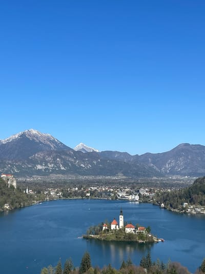 The image is a landscape photograph of Lake Bled in Slovenia. It captures the iconic Bled Island with the Church of the Assumption, its white building and tall spire contrasted by its red roof. The image also includes Lake Bled, the town and the mountains. The sky is blue.