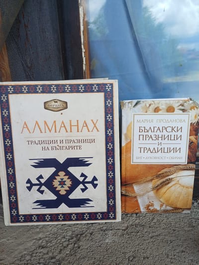 The image displays two books laid side-by-side on a rough, greyish-brown surface, possibly concrete or stucco, with a blurry light blue and dark blue background suggesting a window or protective sheeting.
The upper book, oriented vertically, has a beige and light brown cover. It features an image of a rustic round loaf of bread, several stalks of wheat, and scattered wheat grains on a white cloth. A white, gold-bordered label on the cover reads in Bulgarian: "МАРИЯ ПРОДАНОВА" (Maria Prodanova) as the author, and "БЪЛГАРСКИ ПРАЗНИЦИ И ТРАДИЦИИ" (Bulgarian Holidays and Traditions) as the title. Below the main title, smaller text lists keywords: "БИТ • ДУХОВНОСТ • ОБИЧАИ" (Daily life • Spirituality • Customs).
The lower book has a white cover with a striking dark blue, red, and beige geometric pattern, reminiscent of traditional Bulgarian folk art or textiles, forming a border and a large central emblem. A small brown rectangular logo labeled "ΦΕΡМАТА" (Fermata) is on the left. The title, "АЛМАНАХ" (Almanac), is written vertically in prominent orange letters. To its right, the subtitle, also in Bulgarian, states: "ТРАДИЦИИ И ПРАЗНИЦИ НА БЪЛГАРИТЕ" (Traditions and Holidays of the Bulgarians).
Both books appear to be cultural resources focused on Bulgarian customs and celebrations, aligning with the given location context of Bregovo, Bulgaria. The natural light suggests the photo was taken during the day. There are no people visible, and no specific activity is taking place.