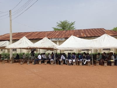 The image depicts a gathering of people under white tents in front of a building. The building is marked "JOLLY NYAN HALL". It is likely this event is taking place in Rumde Waziri, Nigeria, at the latitude and longitude provided. The individuals are predominantly men, many dressed in formal attire, sitting on benches arranged in rows under three tents. They are participating in a social gathering. The ground is dry and dusty. Overhead power lines run from a utility pole across the scene. There are multiple windows visible on the building in the background. A row of trees partially obscures the back of the building.
