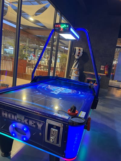 The image shows an air hockey table, illuminated with blue and green lights, positioned indoors near a large window. Outside the window, one can see a multi-story building, possibly a shopping mall, with a distinctive awning featuring triangular fabric shades. A sign for "BURGER" is visible on one of the lower-level storefronts. There are people visible inside the building across from the window. A fire extinguisher is mounted on a wall in the background. The table features a coin slot and the word "HOCKEY" written vertically down the side. Given the architectural style and commercial presence, it suggests the scene is located in a modern entertainment area or shopping center within Mechouar Kasbah, Morocco.