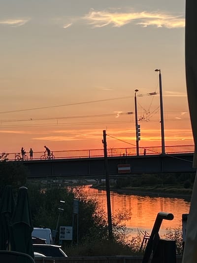 A sunset scene over a body of water, likely a river or canal, in Mannheim, Germany. A bridge crosses the water, with silhouetted figures on it, some of whom are on bicycles. The sky is a gradient of orange and yellow, with wispy clouds. The water reflects the colors of the sunset. The foreground shows trees and some covered umbrellas, suggesting an outdoor seating area, possibly a restaurant or cafe. Electrical infrastructure, including poles and wires, is visible above and on the bridge. A sign with red and white stripes is affixed to the side of the bridge. The overall impression is one of a peaceful, late evening atmosphere.