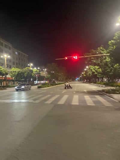 This image depicts a nighttime urban street scene at an intersection in Dali City, China. The multi-lane asphalt road is illuminated by numerous streetlights and features a prominent pedestrian zebra crossing in the foreground. Overhead, a red traffic light displays a digital countdown of '43'.

On the left side of the roadway, a light-colored sedan is positioned in the left lane, with its headlights activated. To the right of the center line, an electric scooter or small motorcycle, occupied by two individuals, is stationary on the road surface, near the crosswalk markings, facing away from the camera. Multi-story buildings with visible illuminated windows line the left side of the street. Trees and planted medians border both sides of the thoroughfare, separating traffic lanes and sidewalks. The sky is dark, indicating night conditions.