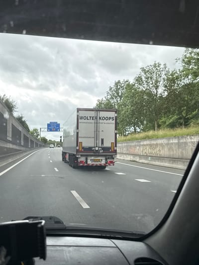 The image shows a view from inside a vehicle on a highway. A white semi-trailer truck with "WOLTER KOOPS INTERNATIONAL TRANSPORT & LOGISTICS" written on its side is in the right lane. The truck has a license plate that is partially visible and appears to read "35-BTZ-3". A road sign is visible in the distance with destinations including "Hoogeveen", "Utrecht", and "Meppel-Zuid", indicating the general location is near Meppel, Netherlands. The highway has multiple lanes and is bordered by a concrete barrier on the left and a grassy embankment with trees on the right. The sky is overcast with clouds. The interior of the vehicle, including the dashboard and windshield, is visible in the foreground.
