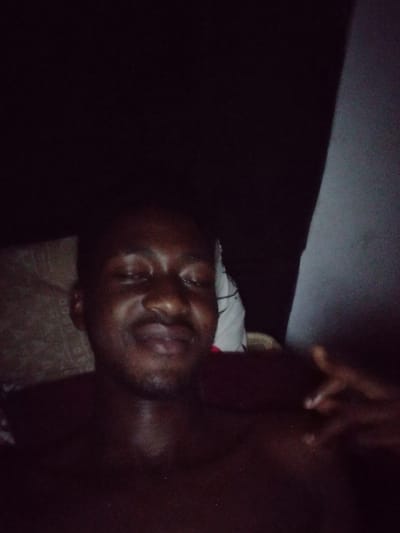 The image shows a male individual, appearing to be an adult with a dark complexion and minimal facial hair (stubble), lying down in a low-light indoor setting. He is positioned facing slightly upwards and towards the viewer, with his eyes closed or squinting and a subtle smile or smirk on his lips, indicating a relaxed or contented state. He appears shirtless or is wearing a very dark garment that blends with the shadows, exposing his upper torso. To the left side of the frame, behind his head, a white object, possibly a pillow or folded fabric with faint dark markings, is visible. His left hand is partially visible and blurred on the right side of the image, with fingers gently splayed. The background on the left is a dark, textured surface, while the right side features a distinct diagonal lighter grey surface, possibly a wall or corner. The scene is presumed to be located in the city of Ogbogbo, Nigeria, as instructed, although no specific visual elements within the image itself confirm this geographical context.