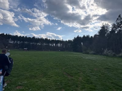 The image shows a grassy field in Topolovgrad, Bulgaria, bordered by a dense line of tall, dark green trees at the horizon. The sky is partly cloudy, with patches of blue visible between the white and gray clouds. In the distance, a soccer goal is visible on the field. In the foreground, a person is partially visible on the left side of the image. The person is wearing dark clothing and appears to be carrying items, including what looks like a tennis racket or similar object. The field has small white flowers scattered throughout the grass. There are also buildings in the background, in the center to upper left.