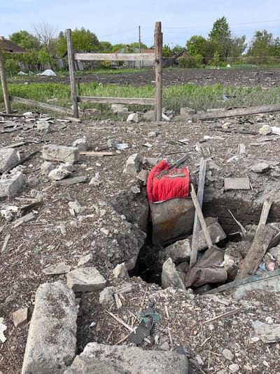 The image shows a crater in the ground, likely caused by an explosion. A red blanket lies in the crater, partially covered by a metal object. The surrounding area is littered with debris, including wood and concrete. The scene is located in the city of Bilopillia, Ukraine, at the coordinates 51.139 latitude and 34.285 longitude. There are no people visible in the image. There is a wooden fence in the background with green vegetation surrounding the crater. The scene is consistent with damage caused by an explosion.