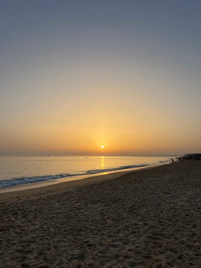 The image displays a wide-angle view of a beach at a low sun angle, consistent with sunrise or sunset, located near Almancil, Portugal. The sky exhibits a gradient, transitioning from a light blue-grey at the zenith to a bright orange-yellow at the horizon. A single, small, dark avian form is visible in the upper central sky.

The sun, a distinct orange-yellow disc, is positioned just above the horizon, centrally located in the frame. It casts a direct, reflective light path across the calm ocean surface towards the viewer. The ocean edge features gentle wave breaks with visible foam.

The foreground and midground are composed of light brown sand, textured with minor undulations and indistinct traces of human passage. The beach extends longitudinally into the distance on the right. In the far distance on the water, multiple small, dark, unidentifiable objects, possibly maritime vessels or markers, are present.

The far right section of the beach contains multiple human figures, rendered as dark silhouettes against the ambient light. These individuals are sparsely distributed along the sand and near the water's edge. Their specific actions or interactions are not discernible due to distance and backlighting.