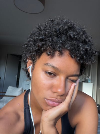 A close-up, portrait-oriented image shows a person with dark, tightly coiled curly hair and brown skin, captured indoors in Champs-sur-Marne, France. The subject's body is oriented horizontally across the frame, with their head resting on their left arm, while their face is angled towards the camera. They look directly at the viewer with a relaxed, contemplative expression. Their left hand (viewer's right) gently cups their temple and cheek.

The person is wearing a dark tank top or sports bra. White wired earphones are plugged into their left ear, which is adorned with a small hoop earring and another small, possibly diamante, earring in the cartilage. Their skin appears clear and dewy, and their lips are slightly parted. A small blemish is visible on their right collarbone area (viewer's left).

The background is softly blurred, indicating an indoor setting with natural light coming from the right, creating highlights on the subject's hair and skin. On the far left, a light-colored, circular object (possibly a lamp or part of furniture) is subtly visible. To the right, a blurry white structure, possibly shelving or a counter, and a white garment with faint, illegible text are visible. The overall scene suggests a casual, personal moment.