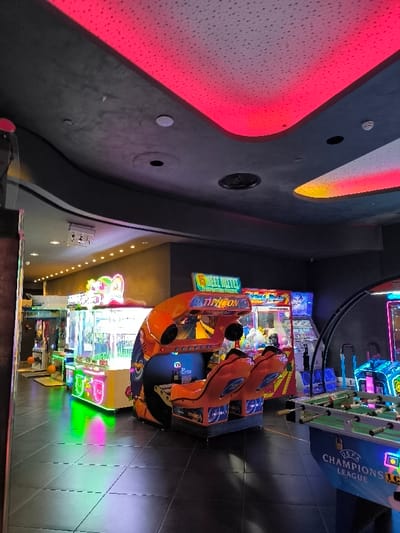 This image captures the interior of a modern arcade or entertainment center, likely located in Agios Dimitrios, Greece. The scene is brightly lit by the vibrant displays of numerous arcade games, set against a backdrop of dark walls and ceiling.

The main subjects are various arcade machines. In the foreground and center is a prominent orange racing or driving simulator game, featuring two seats and a screen, with the word "TYPHOON" clearly visible on its structure. Above this game, partially obscured, is text that appears to read "5 WHEEL HELL". To the left and further into the background, several claw machines and other prize games glow with multi-colored lights, casting reflections on the dark, tiled floor. One such claw machine has "SWEET HOLLY" partially visible on its marquee. To the right, part of a foosball table is visible, prominently displaying the "UEFA CHAMPIONS LEAGUE" logo on its side, suggesting a sports-themed game.

No people are present in the image, indicating either an early opening, a late closing, or a quiet moment at the venue. The overall ambiance is one of leisure and entertainment.

Notable details include the ceiling, which is a key architectural feature. It is dark gray with recessed sections that are illuminated by strip lighting. A significant portion of the ceiling on the left and center glows with a vibrant red light, while a section on the right emits a warm yellow-orange hue. These LED lights create a dynamic and inviting atmosphere typical of such entertainment venues. The floor consists of dark, rectangular tiles, reflecting the bright lights from the games. The time of day cannot be determined due to the indoor, artificial lighting, but the dim ambient light suggests an evening setting. The overall impression is a clean, modern, and engaging space designed for gaming and fun.