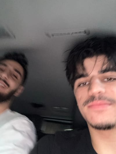 Two adult males are depicted inside what appears to be a vehicle interior. The male on the right occupies the majority of the right frame, featuring dark, disheveled hair, brown eyes, a mustache, and a short beard. He wears a dark-colored shirt and looks directly towards the camera with a neutral expression. The male on the left is partially visible, positioned further back and to the left, with dark hair, a beard, and a light-colored shirt. He is smiling and also appears to be looking towards the camera. The vehicle's headliner is visible in the upper portion of the frame, alongside what may be an overhead console or light fixture. The lighting is subdued. The specific location within the city of Fort Lee, United States, cannot be determined from the visual evidence presented in the image.