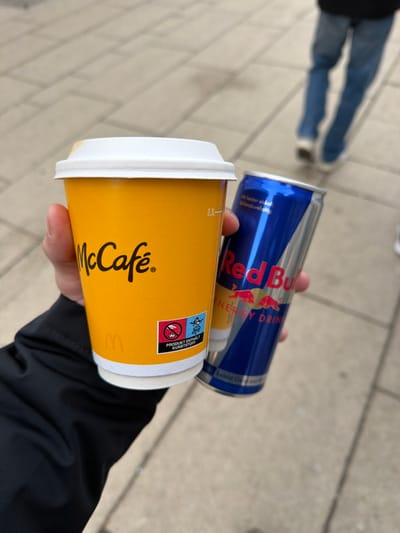 The image presents a close-up, eye-level view of a hand holding two distinct beverage containers on a paved surface, purportedly located in Sankt Pölten, Austria. A light-skinned human hand, partially covered by a black jacket or sleeve, grasps a yellow McCafé disposable cup with a white lid in the left fingers, and a blue and silver Red Bull energy drink can in the right fingers. The McCafé cup features the brand's logo in black, a faint McDonald's 'M' emblem near the bottom, and a sticker with German text "PRODUKT ENTHÄLT KUNSTSTOFF" (Product Contains Plastic) and a "no littering" pictogram. A "0.3L" volume indicator is visible on the cup. The Red Bull can displays its prominent branding, including "Red Bull ENERGY DRINK" and two red bull motifs. In the blurred background, the lower half of another individual is visible, wearing blue denim jeans and light-colored shoes, walking away from the camera. The ground is a light-toned paved surface, consisting of rectangular or square tiles. The ambient lighting suggests an outdoor, possibly overcast, daytime environment.
