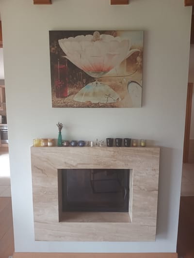 The image shows a living room with a fireplace. Above the fireplace, there is a painting of a large pink flower. The fireplace is made of beige marble and has a dark-colored insert. On the mantlepiece, there are several decorative items, including candles, small figurines, and a green vase with dried flowers. To the left of the fireplace, a section of a kitchen with a stove and oven is visible. The room has light-colored walls and wooden flooring. The overall impression is of a well-decorated and tidy living space. This scene is located in the city of Avižieniai, Lithuania.