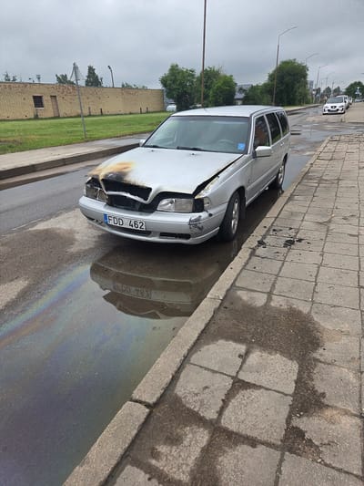 A silver Volvo V70 estate car is parked on the side of a street in Širvintos, Lithuania. The car's hood is damaged and shows signs of fire, with scorched metal and melted plastic visible. The front bumper is also damaged. The car is parked next to a concrete sidewalk, and there is a puddle of water on the road reflecting the car and displaying an oil slick with rainbow-like patterns. The street appears wet, suggesting recent rain or a water spill. In the background, there is a long, low brick building with a small window and a wooden door, and several trees are visible behind it. Other cars are parked further down the street. The sky is overcast. There are no people visible in the image.