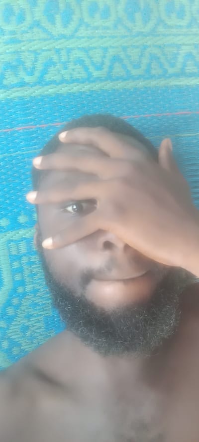 A dark-skinned man with a beard covers the top half of his face with his hand, obscuring his forehead and one eye. The visible eye looks directly at the camera. He appears to be lying down on a blue and green patterned woven mat. The background is blurry, but the mat and the man are the only subjects visible in the scene. There is no further information to indicate that the picture was taken in Bende, Nigeria. The picture does not show any other objects.