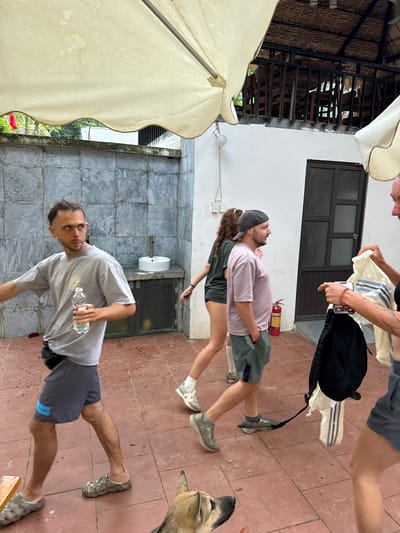An outdoor courtyard in Cổ Lũng Commune, Vietnam, features a group of people and a dog. A man in a grey t-shirt and dark shorts holds a water bottle, walking towards the left. Behind him, a woman in a dark green t-shirt and shorts walks away from the camera. To her right, a man wearing a pink t-shirt and green shorts, with a grey baseball cap, walks forward. A woman on the far right, wearing a grey outfit, is holding a white shirt with blue stripes and a black backpack. A dog with light brown fur is visible in the lower right corner, facing left. The courtyard has a tiled floor and a stone wall. In the background, there is a building with a thatched roof and wooden railings. A fire extinguisher is visible on the wall.