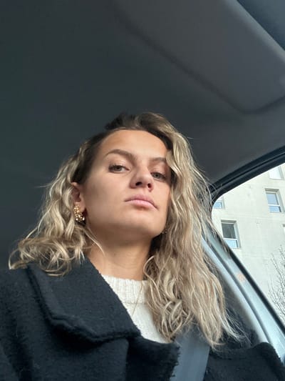 A woman with wavy blonde hair peers at the camera from an angled perspective. She has fair skin, light brown eyes, and pink lips. She wears a white sweater and a black coat. Her earring, a cluster of small gold orbs, is visible on one ear. The background shows the interior of a car, the roof predominantly taking up the space, and an apartment building is seen through the car window.
