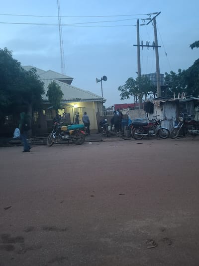 The image depicts a street scene in Karu, Nigeria, likely during the early morning or late evening given the soft, diffused light. The orientation of the image is rotated 90 degrees clockwise.
The primary subjects are several motorcycles parked along a paved road. Some of these motorcycles are carrying cargo, suggesting they are used for transport. A few people are visible near the motorcycles and a building. One person in a green shirt and blue jeans is standing on the side of the road, while others are gathered near the building and motorcycles, possibly loading or unloading goods.
The building behind the motorcycles appears to be a commercial or residential structure with a light-colored, sloped roof. Electrical poles with wires are prominent in the background, indicating an urban or semi-urban environment. The overall scene suggests a typical day in a bustling area where motorcycles serve as a vital mode of transportation. The weather appears to be clear, with a pale blue sky. There is no visible text in the image.