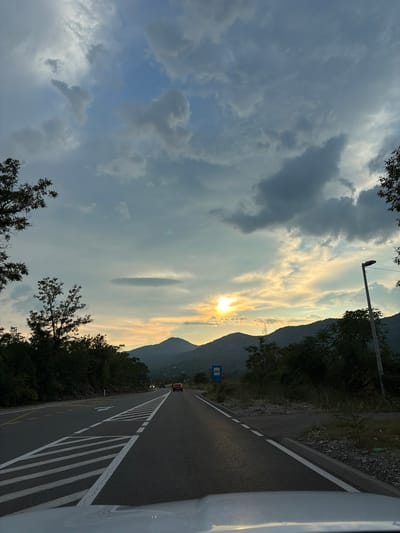 The image shows a road in Barutana, Montenegro, during what appears to be sunset. The sky is filled with a mix of dark and light clouds, with the sun peeking through, casting a warm glow on the horizon. Mountains form the backdrop, covered in dense green foliage. On the right side of the road, there's a streetlamp and some dry grass and rocks. On the left, a dense line of trees lines the road. The road itself is asphalt with clear white markings. There are a few cars on the road, indicating traffic. A blue bus stop sign is visible on the side of the road. The view appears to be from inside a vehicle, possibly a car, judging by the white hood visible at the bottom of the frame.