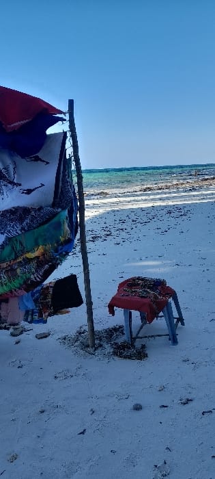 A bright outdoor scene depicts a sandy beach in Ziwa La Ng'ombe ward, Kenya, under a clear blue sky. In the left foreground, a vertical wooden pole, anchored in the sand with surrounding debris, supports a display of multiple patterned fabrics, including items with giraffe and abstract designs, draped over it and partially under a red canopy. Smaller garment-like items are visible hanging lower. To the right, a light blue plastic stool is covered with a red fabric, upon which several smaller, patterned items or crafts are arranged. The midground consists of an expansive, light-colored sandy beach with scattered dark debris, possibly seaweed or small rocks. The background features turquoise-blue ocean water transitioning to a darker blue further out, meeting a clear, light blue sky at the horizon. Shadows are visible on the sand, consistent with direct sunlight. No individuals are clearly discernible in the immediate foreground or midground.