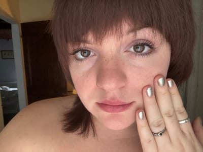 An indoor, eye-level photograph depicts a fair-skinned individual with short, dark reddish-brown hair cut with bangs, directly facing the camera. The individual has brown eyes with visible eyelashes, light pink eyeshadow, a gold nose ring in the left nostril, and light pink lips, with freckles visible on the cheeks. The left bare shoulder is partially exposed. The individual's left hand is positioned with fingers resting against the left cheek. The fingernails are covered in silver glitter polish. A plain silver-toned band adorns the left ring finger, and a textured silver-toned ring with a black circular stone is on the left middle finger. The background is out of focus, showing a dark brown door or wall to the right, and a partially obscured doorway leading into another dimly lit room on the left. Within the distant room, indistinct furniture, possibly a bed, and a framed object on a wall are visible. The setting is an interior space in Montepulciano, Italy.