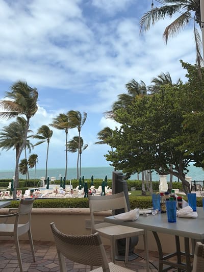 The image captures a serene outdoor scene, likely at a resort or hotel in Key Biscayne, United States of America. The foreground includes a table set with blue cups, white napkins, and various bottles, suggesting a bar or dining area. Wicker chairs are arranged around the table. Beyond the table, a landscaped area with lush greenery and palm trees leads to a view of the ocean. People are lounging on beach chairs under green umbrellas along the waterfront. The sky is partially cloudy, creating a bright and airy atmosphere. The scene depicts a relaxing and luxurious vacation setting.