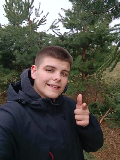 This media file features a cheerful young man, likely in his late teens or early twenties, captured in a selfie outdoors in Krasnyi Mayak, Russia. He has a fair complexion, short, neatly trimmed dark brown hair, and warm brown eyes. A broad smile lights up his face, revealing his teeth, and his gaze is directed straight at the viewer. He wears a practical, dark-colored (possibly black or navy blue) hooded winter jacket, equipped with drawstrings around the hood and a central zipper, suggesting protection against cool or cold weather.
The man’s right hand is prominent in the foreground, with his index finger pointing directly towards the camera, conveying a sense of direct engagement or invitation. The background is a natural landscape dominated by numerous healthy green pine trees or conifers, their needles dense and vibrant. Patches of dry, brown grass and fallen branches are visible on the ground beneath the trees. The lighting appears soft and diffused, characteristic of an overcast day, with no harsh shadows present, and a bright, featureless sky visible in the upper sections of the image. The cool attire and the type of foliage are consistent with a late autumn, winter, or early spring setting. No visible text is present in the image. The overall impression is one of friendliness and candidness.