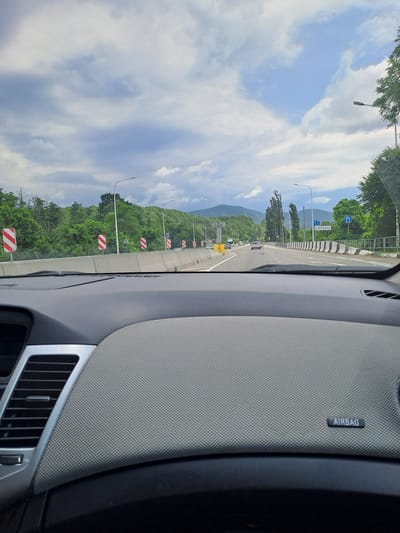 This media file depicts a view from inside a car traveling on a highway in a mountainous, forested region. The image is oriented vertically.

**Subjects:** The primary subjects are the road, the car's interior, and the natural landscape. The car's dashboard, including an air vent and the "AIRBAG" label, is visible on the right side. The road stretches forward, with other vehicles ahead. The left side of the image showcases a lush green mountainside covered in trees. The sky above is partly cloudy, with white and blue patches.

**Activity or event:** The image captures a moment during a car journey. The perspective suggests the vehicle is in motion, moving along the highway.

**Notable details:**
*   **Time of day/Weather:** The bright sky with scattered clouds indicates it is daytime, likely late morning or afternoon. The weather appears to be fair.
*   **Location cues:** There are road signs visible. One sign has blue text and arrows, and another, which is partially obscured, appears to be a directional sign with Cyrillic text that reads "р. ДЕСАНЬ" (R. DESAN'). Road markings and barrier delineators with red and white diagonal stripes are present along the side of the road, indicating a curve or hazard. Streetlights are also visible. The mountainous and heavily forested terrain suggests a rural or semi-rural location. The presence of the "AIRBAG" label points to a modern passenger vehicle.
*   **Emotions:** The image does not convey any explicit emotions.

**Visible text:** The word "AIRBAG" is clearly visible on a label inside the car. A road sign displays Cyrillic text that appears to read "р. ДЕСАНЬ".